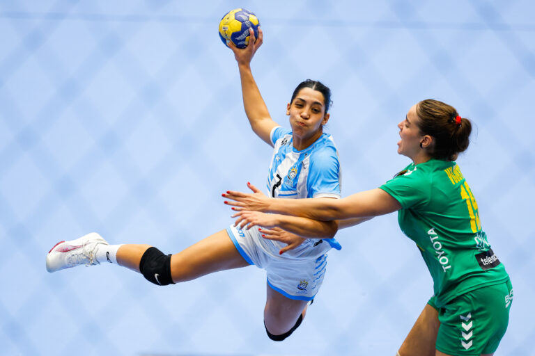 An athlete in a light blue uniform leaps to catch a handball while being defended by an opponent in a green uniform.